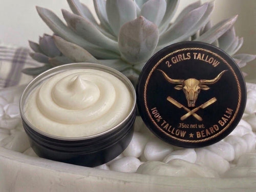 Open black tin of smooth, creamy off-white beard balm, with its black lid propped beside it. The lid shows a gold longhorn skull over crossed straight razors inside a rope border, with the words "2 Girls Tallow," "100% Tallow Beard Balm," and ".75oz net wt." The tin sits on white pebbles in front of a pale green succulent.