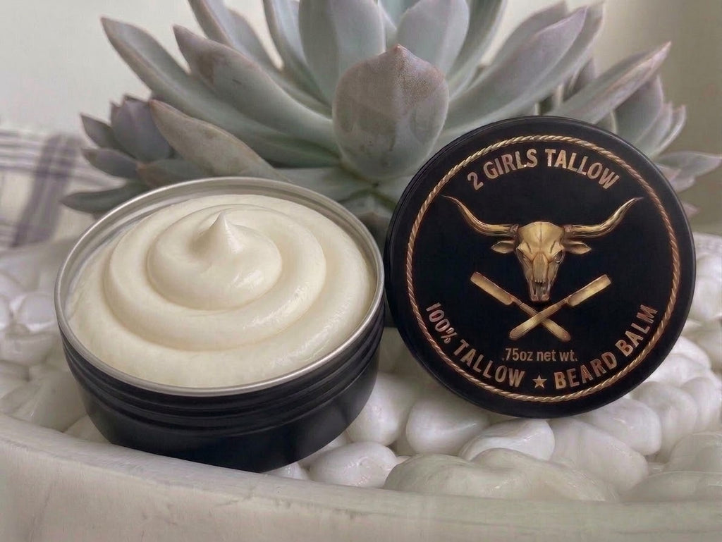 Open black tin of smooth, creamy off-white beard balm, with its black lid propped beside it. The lid shows a gold longhorn skull over crossed straight razors inside a rope border, with the words "2 Girls Tallow," "100% Tallow Beard Balm," and ".75oz net wt." The tin sits on white pebbles in front of a pale green succulent.