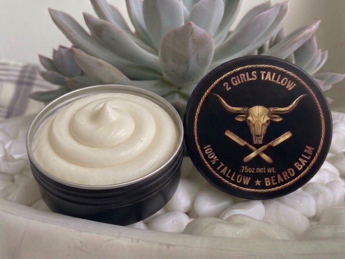 Open black tin of smooth, creamy off-white beard balm, with its black lid propped beside it. The lid shows a gold longhorn skull over crossed straight razors inside a rope border, with the words "2 Girls Tallow," "100% Tallow Beard Balm," and ".75oz net wt." The tin sits on white pebbles in front of a pale green succulent.