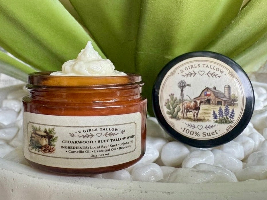 Open amber glass sample jar of 2 Girls Tallow Cedarwood Suet Tallow Whip, topped with a swirl of white whipped tallow. The black lid rests beside it, showing a farm scene with a longhorn, a barn with a Texas flag, a windmill and bluebonnets, and the words "100% Suet." The jar's label lists local beef suet, jojoba oil, camellia oil, essential oil and beeswax. The jar and lid sit on white pebbles in front of green leaves.