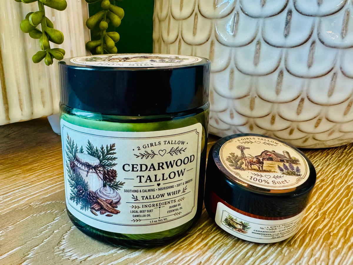 Large green glass jar of 2 Girls Tallow Cedarwood Tallow Whip with a black lid and a cream label showing cedar branches and pine cones, next to a smaller sample jar with a dark lid showing a farm scene and the words "100% Suet." Both sit on a light wooden table in front of a white ceramic planter with scalloped pattern and trailing green plants.