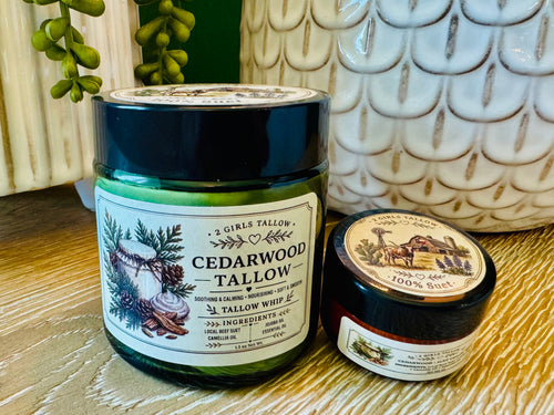 Large green glass jar of 2 Girls Tallow Cedarwood Tallow Whip with a black lid and a cream label showing cedar branches and pine cones, next to a smaller sample jar with a dark lid showing a farm scene and the words "100% Suet." Both sit on a light wooden table in front of a white ceramic planter with scalloped pattern and trailing green plants.