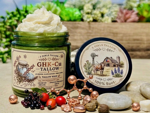 Open green glass jar of 2 Girls Tallow GHK-Cu Tallow Whip, topped with a tall swirl of white whipped tallow. Its black lid rests beside it, showing a farm scene with a longhorn, barn, windmill and bluebonnets, and the words "100% Suet." In front are copper spheres marked "Cu²⁺" joined into a molecule model, raw copper nuggets, red rose hips, dark berries and smooth stones, on a rustic wooden surface with greenery and succulents behind.