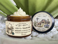 Open amber glass sample jar of 2 Girls Tallow GHK-Cu Tallow Whip, topped with a swirl of white whipped tallow. The label lists local beef suet, jojoba oil, camellia oil, copper tripeptide-1 and unscented, .25oz net wt. The black lid rests beside it, showing a farm scene with a longhorn, a barn with a Texas flag and bluebonnets, and the words "100% Suet." Jar and lid sit on white pebbles in front of green leaves.