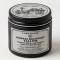 Black glass jar with a black screw lid, on a light linen background. The silver lid label shows an engraved-style farm scene with a longhorn, barn, windmill and cactus, ringed by "2 Girls Tallow" and "100% Suet." The front label reads "2 Girls Tallow, Herbal Recovery Tallow, Hydrating Muscle & Joint Recovery, 2 Girls Artisanal Suet Whip," with ingredients beef suet, jojoba and camellia oil, squalane, ceramide NP, wintergreen, camphor, peppermint, and various essential oils, flower oils and extracts. 1.5 oz 
