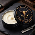 Open black tin of creamy off-white men's suet hydration whip, its lid propped beside it showing a gold longhorn skull and crossed straight razors inside a rope border. Lid text reads "2 Girls Tallow, Men's Suet Hydration Whip, .75oz net wt." The tin sits on a dark slate board next to a straight razor, with a leather wallet in the background.