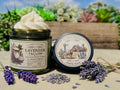 Open green glass jar of 2 Girls Tallow Lavender Tallow Whip, topped with a tall swirl of white whipped tallow, with lavender sprigs on its label. The black lid rests beside it, showing a farm scene and the words "100% Suet." A bundle of fresh lavender tied with twine, loose dried lavender buds and scattered blossoms surround them on a tabletop, with a wooden planter of succulents and blue sky behind.