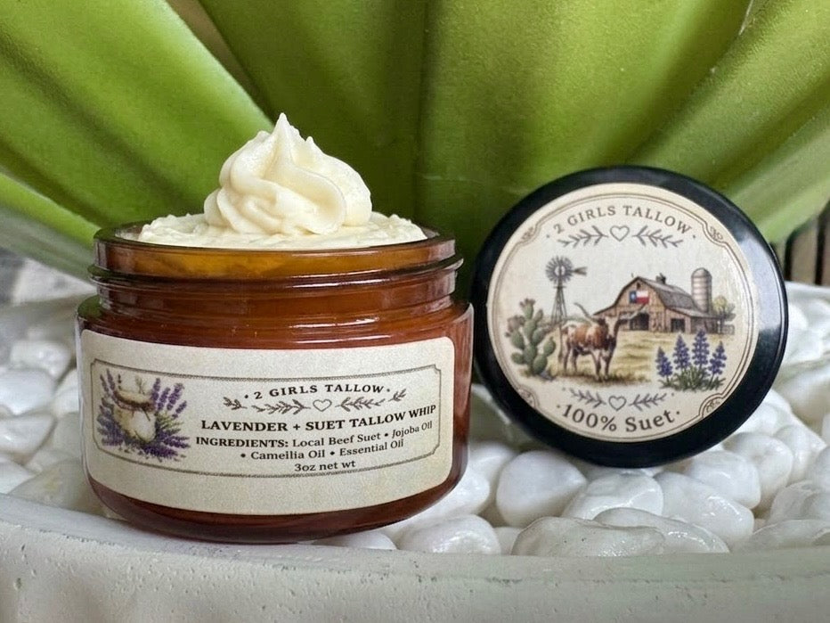 Open amber glass sample jar of 2 Girls Tallow Lavender Suet Tallow Whip, topped with a swirl of white whipped tallow. The label shows lavender sprigs and lists local beef suet, jojoba oil, camellia oil and essential oil. The black lid rests beside it, showing a farm scene with a longhorn, a barn with a Texas flag and bluebonnets, and the words "100% Suet." Jar and lid sit on white pebbles in front of green leaves.