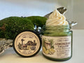 Open green glass jar of 2 Girls Tallow Lemongrass Tallow Whip, topped with a tall swirl of white whipped tallow. The label shows a small jar with lemongrass stalks and lemon wedges. The black lid leans beside it, showing a farm scene with a longhorn, a barn with a Texas flag and bluebonnets, and the words "100% Suet." Green moss and silvery decorative pieces sit behind, on a white shelf.