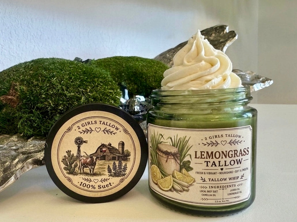 Open green glass jar of 2 Girls Tallow Lemongrass Tallow Whip, topped with a tall swirl of white whipped tallow. The label shows a small jar with lemongrass stalks and lemon wedges. The black lid leans beside it, showing a farm scene with a longhorn, a barn with a Texas flag and bluebonnets, and the words "100% Suet." Green moss and silvery decorative pieces sit behind, on a white shelf.