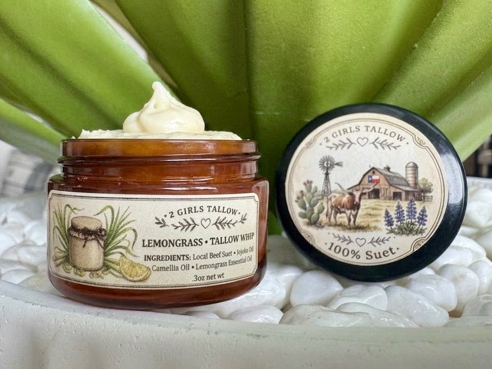 Open amber glass sample jar of 2 Girls Tallow Lemongrass Tallow Whip, topped with a swirl of pale cream whipped tallow. The label shows a lemongrass illustration and lists local beef suet, jojoba oil, camellia oil and lemongrass essential oil. The black lid rests beside it, showing a farm scene with a longhorn, a barn with a Texas flag and bluebonnets, and the words "100% Suet." Jar and lid sit on white pebbles in front of green leaves.