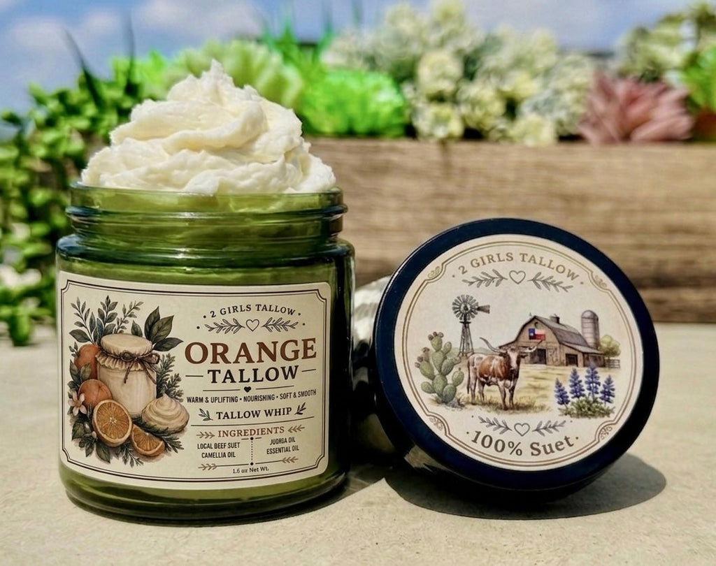 Open green glass jar of 2 Girls Tallow Orange Tallow Whip, topped with a tall swirl of white whipped tallow. The label shows orange slices and leaves. The black lid leans beside it, showing a farm scene with a longhorn, a barn with a Texas flag and bluebonnets, and the words "100% Suet." The jar and lid sit on a stone tabletop in front of a wooden planter of succulents, with blue sky behind.