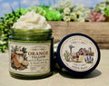 Open green glass jar of 2 Girls Tallow Orange Tallow Whip, topped with a tall swirl of white whipped tallow. The label shows orange slices and leaves. The black lid leans beside it, showing a farm scene with a longhorn, a barn with a Texas flag and bluebonnets, and the words "100% Suet." The jar and lid sit on a stone tabletop in front of a wooden planter of succulents, with blue sky behind.