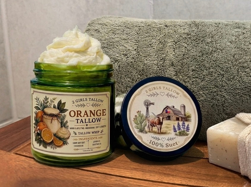 Open green glass jar of 2 Girls Tallow Orange Tallow Whip, topped with a tall swirl of white whipped tallow, with orange slices on its label. The black lid leans beside it, showing a farm scene and the words "100% Suet." A folded gray-green towel and a cream-colored bar of soap wrapped in a cloth band sit behind and beside them on a wooden surface.
