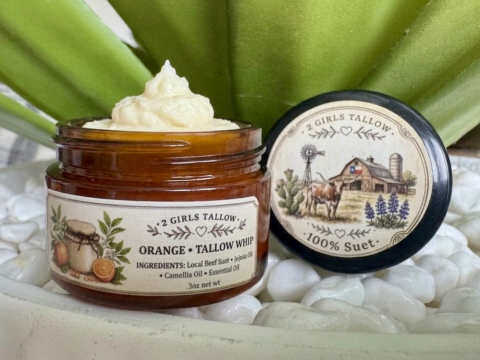 An amber glass jar of Orange + Tallow Whip by 2 Girls Tallow, topped with a swirl of white cream, shown next to its black-lidded jar on white pebbles with green leaves in the background.