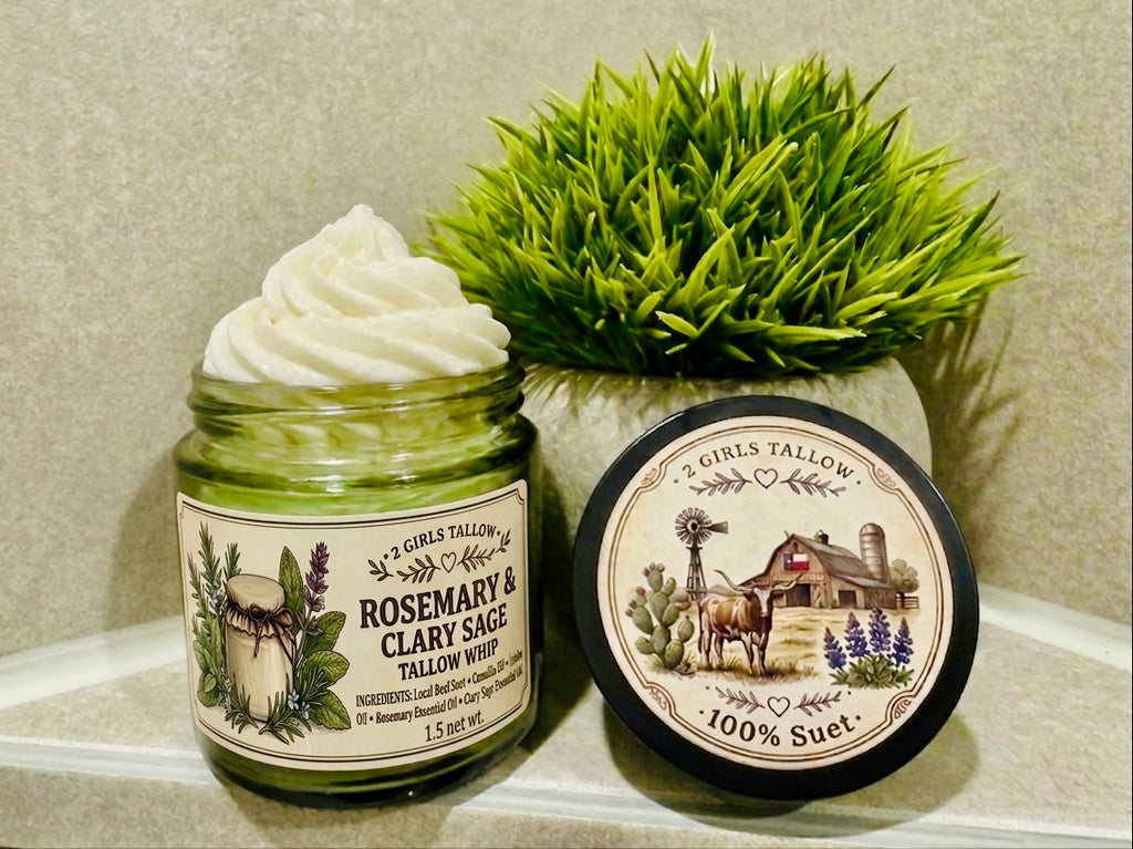 Open green glass jar of 2 Girls Tallow Rosemary & Clary Sage Tallow Whip, topped with a tall swirl of white whipped tallow, with rosemary and sage sprigs on its label. The black lid rests beside it, showing a farm scene with a longhorn, a barn with a Texas flag and bluebonnets, and the words "100% Suet." A potted green plant sits behind them, on a pale shelf against a gray-green tiled wall.