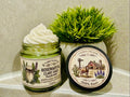 Open green glass jar of 2 Girls Tallow Rosemary & Clary Sage Tallow Whip, topped with a tall swirl of white whipped tallow, with rosemary and sage sprigs on its label. The black lid rests beside it, showing a farm scene with a longhorn, a barn with a Texas flag and bluebonnets, and the words "100% Suet." A potted green plant sits behind them, on a pale shelf against a gray-green tiled wall.