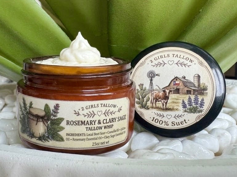 Open amber glass sample jar of 2 Girls Tallow Rosemary & Clary Sage Tallow Whip, topped with a swirl of white whipped tallow. The label shows rosemary and sage sprigs. The black lid rests beside it, showing a farm scene with a longhorn, a barn with a Texas flag and bluebonnets, and the words "100% Suet." Jar and lid sit on white pebbles in front of green leaves.