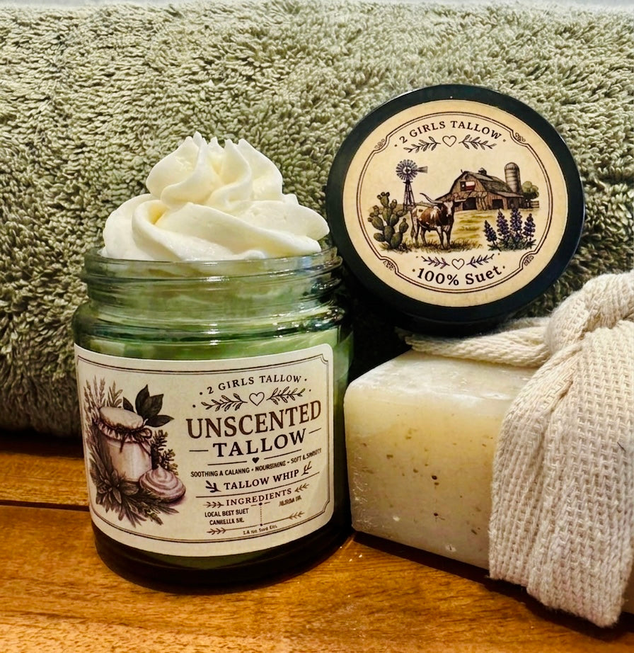 Open green glass jar of 2 Girls Tallow Unscented Tallow Whip, topped with a tall swirl of white whipped tallow. The black lid leans beside it, showing a farm scene with a longhorn, barn and windmill, and the words "100% Suet." A folded gray-green towel and a speckled cream bar of soap in a woven cloth wrap sit behind them on a wooden surface.