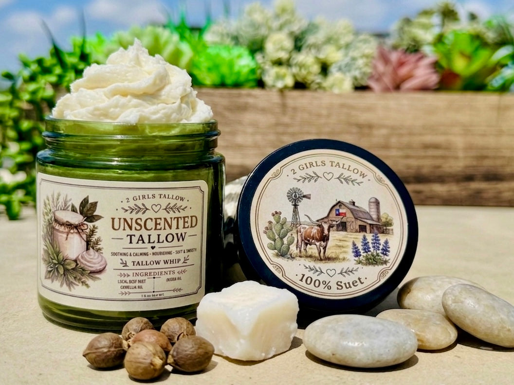Open green glass jar of 2 Girls Tallow Unscented Tallow Whip, topped with a tall swirl of white whipped tallow. The label reads "Unscented Tallow, Tallow Whip." The black lid rests beside it, showing a farm scene with a longhorn, a barn with a Texas flag, a windmill and bluebonnets, and the words "100% Suet." In front are a cube of white tallow, smooth stones and brown nuts, on a tabletop against a blurred garden of succulents.