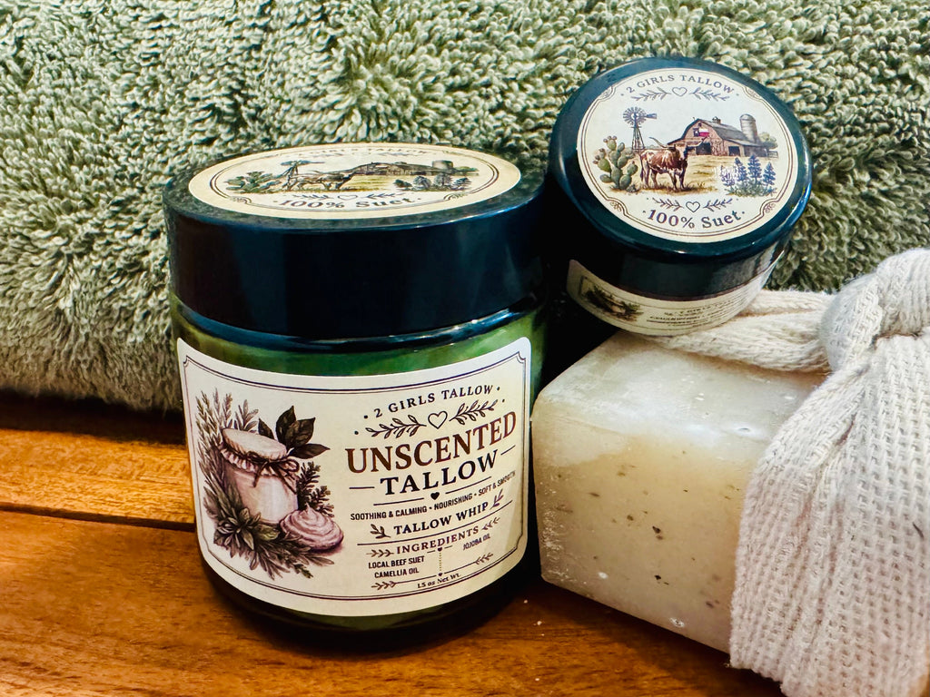 Large closed green glass jar of 2 Girls Tallow Unscented Tallow Whip, with a farm-scene "100% Suet" label on its black lid, next to a smaller sample jar with a matching lid. A speckled cream bar of soap wrapped in woven cloth sits beside them, with a green towel behind, on a wooden surface.