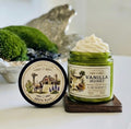 Open green glass jar of 2 Girls Tallow Vanilla Honey Suet Tallow Whip, topped with a tall swirl of white whipped tallow flecked with vanilla, on a dark wooden coaster. The label shows vanilla flowers and a honey dipper. The black lid leans beside it, showing a farm scene with a barn, windmill, cactus, bluebonnets and a grazing animal, and the words "100% Suet." Green moss and a silvery decorative piece sit behind, on a white surface.