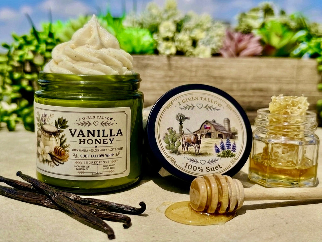Open green glass jar of 2 Girls Tallow Vanilla Honey Suet Tallow Whip, topped with a tall swirl of white whipped tallow flecked with vanilla. The label shows vanilla flowers and a honey dipper. The black lid rests beside it, showing a farm scene with a longhorn, a barn with a Texas flag, a windmill and bluebonnets, and the words "100% Suet." A small glass jar of honey with honeycomb, a wooden honey dipper resting in a pool of honey, and two vanilla beans sit in front, with a blurred planter of succulents be