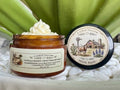 Open amber glass sample jar of 2 Girls Tallow Vanilla Honey Suet Tallow Whip, topped with a swirl of white whipped tallow. The label shows vanilla and honeycomb illustrations and lists local beef suet, local honey, camellia oil and essential oil. The black lid rests beside it, showing a farm scene with a longhorn, a barn with a Texas flag and bluebonnets, and the words "100% Suet." Jar and lid sit on white pebbles in front of green leaves.